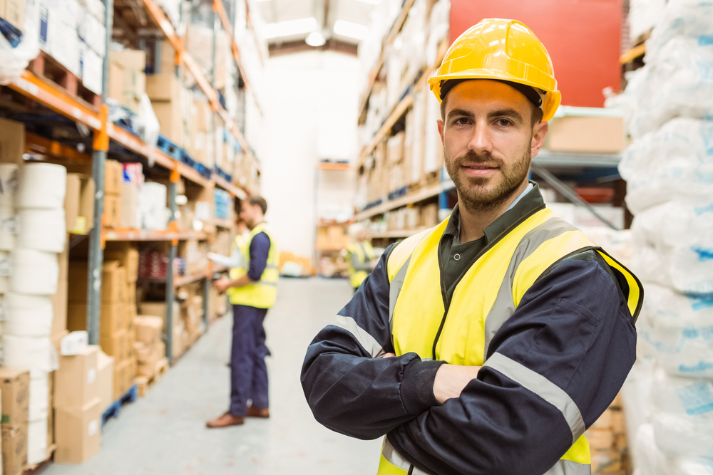 Smiling worker wearing yellow vest with arms crossed in a large warehouse Smiling worker wearing yellow vest with arms crossed in a large warehouse